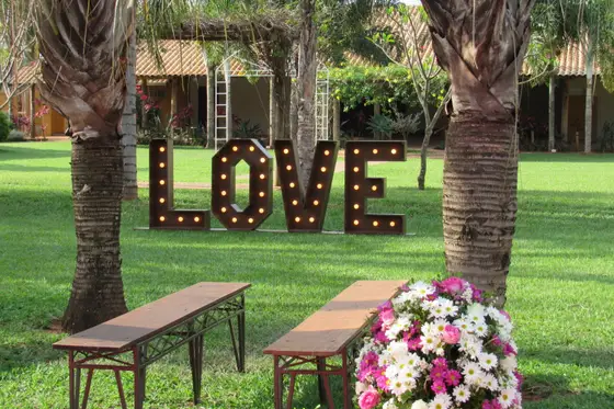 Letreiro luminoso “LOVE” entre palmeiras com banco de madeira e flores