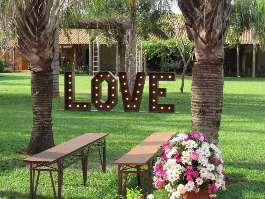 Letreiro luminoso “LOVE” entre palmeiras com banco de madeira e flores